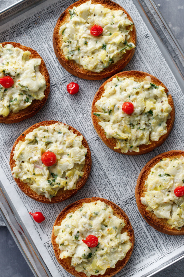 Overhead view of Artichoke Crostini on a baking sheet lined with newspaper-print parchment paper