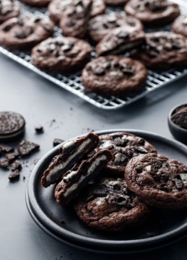 Cream-Stuffed Chocolate Cookies 'n Cream Cookies on a plate, one cut in half to show the cream stuffing, with a baking rack and crumbled cookies in the background.