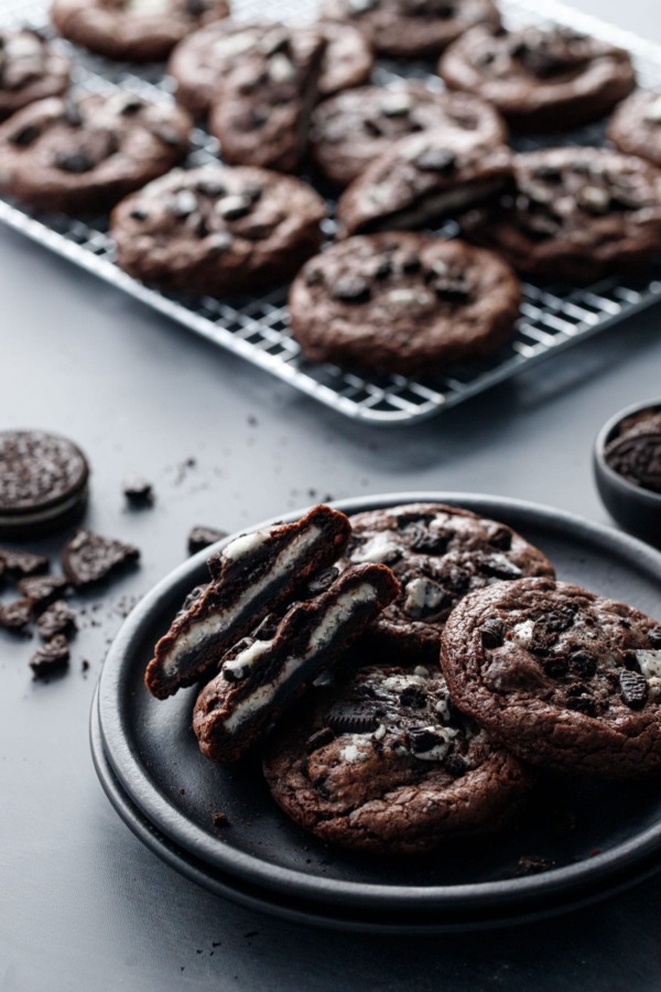 Cream-Stuffed Chocolate Cookies 'n Cream Cookies on a plate, one cut in half to show the cream stuffing, with a baking rack and crumbled cookies in the background.