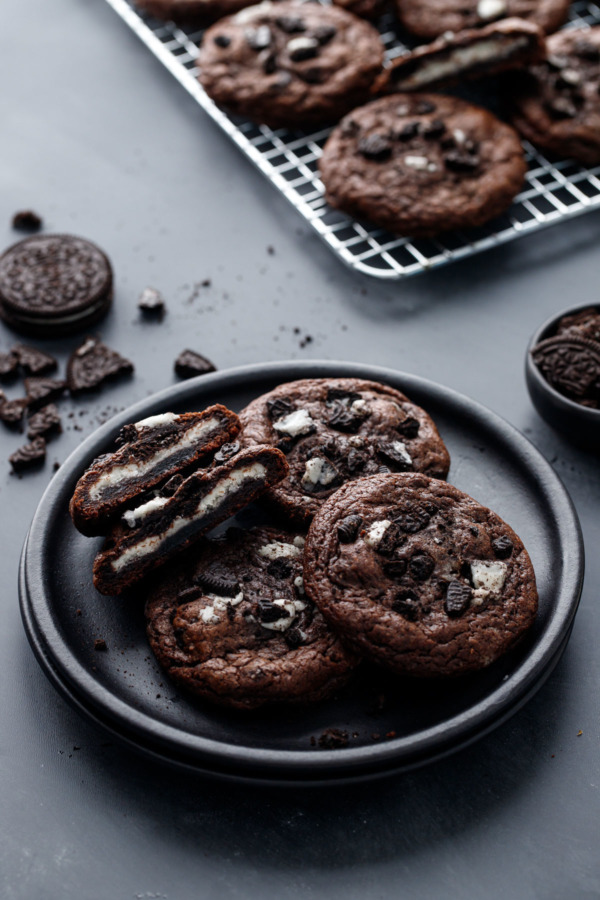 Cream-Stuffed Chocolate Cookies 'n Cream Cookies on a plate, one cut in half to show the cream stuffing, with a baking rack and crumbled cookies in the background.