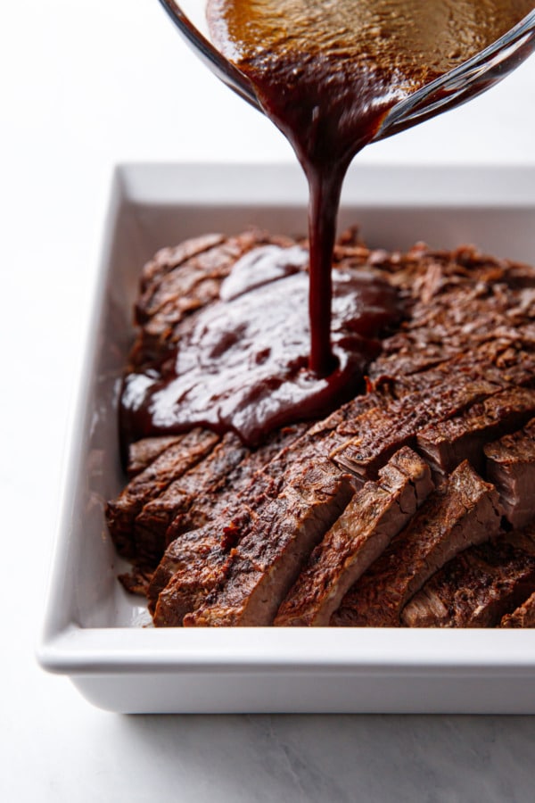 Slow cooker beef brisket sliced and arranged in a white baking dish, sauce pouring over top.