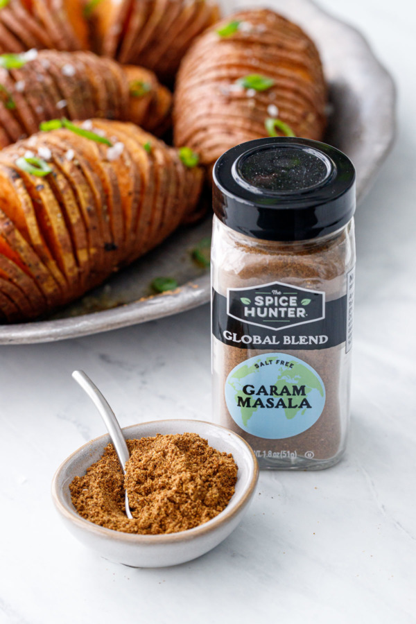 Bottle of The Spice Hunter Garam Masala Blend with a bowl of spice and a tiny spoon; the finished Spiced Hasselback Sweet Potatoes in the background.