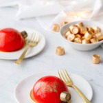 Two red mirror-glazed cakes decorated to look like Christmas ornaments, on white plates with gold forks and fairy lights in the background.