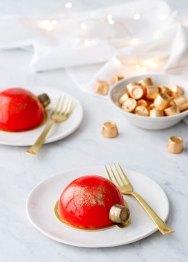 Two red mirror-glazed cakes decorated to look like Christmas ornaments, on white plates with gold forks and fairy lights in the background.