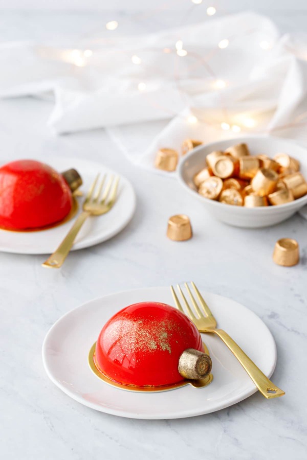 Two red mirror-glazed cakes decorated to look like Christmas ornaments, on white plates with gold forks and fairy lights in the background. 