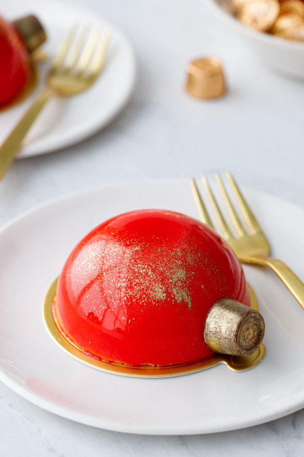 A red mirror-glazed half dome cake with a gold dusted Rolo on the side to look like a Christmas ornament