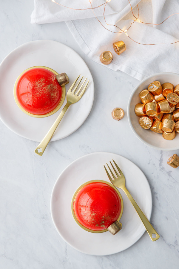 Overhead view of red mirror-glazed entremet cakes decorated to look like Christmas ornaments, with gold forks and a bowl of Rolo candies on the side