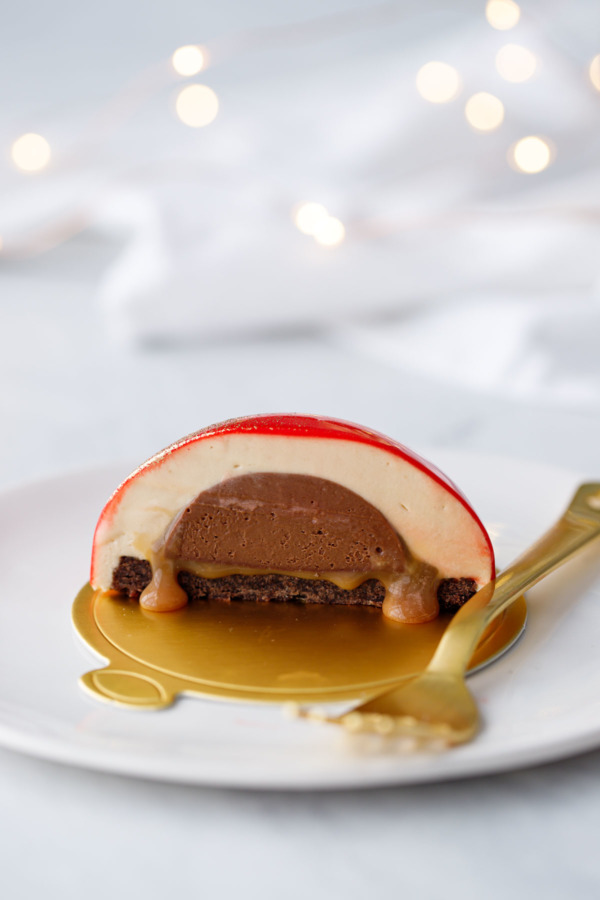 Cross section of an entremet cake with caramel oozing out; on a white plate with a gold fork and fairy lights in the background.