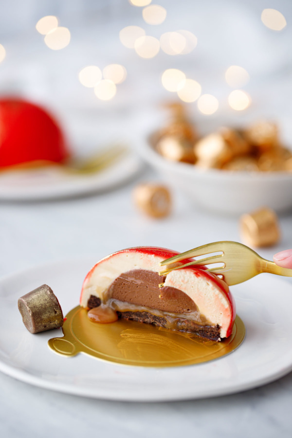 A gold fork cutting in to an entremet cake, on a white plate with a bowl of Rolos and fairy lights in the background