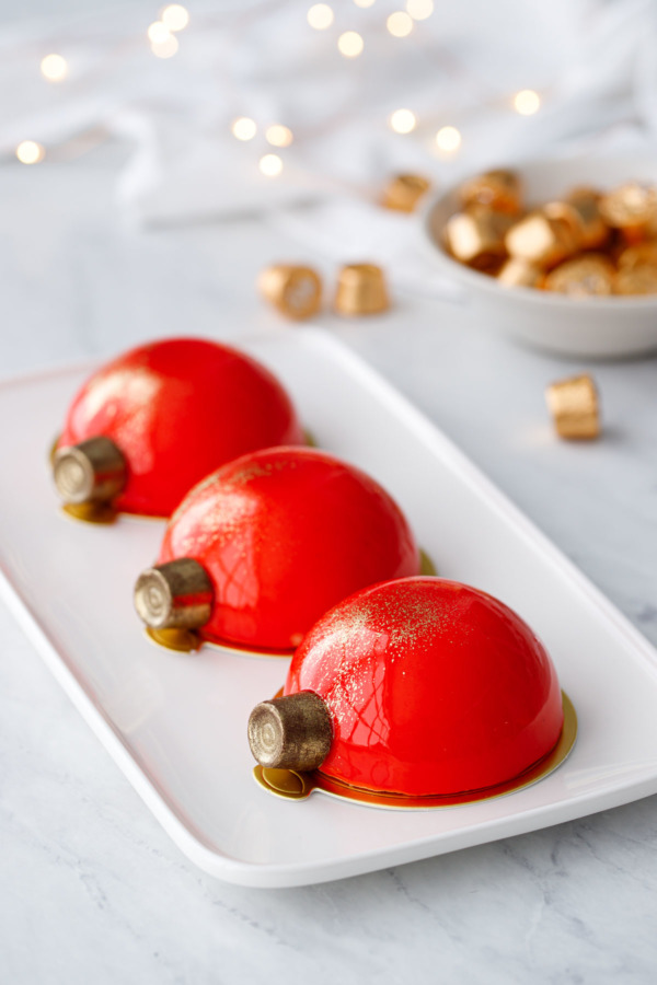 Three red mirror-glazed Christmas ornament cakes on a white rectangle plate with a bowl of Rolo candies and fairy lights in the background.