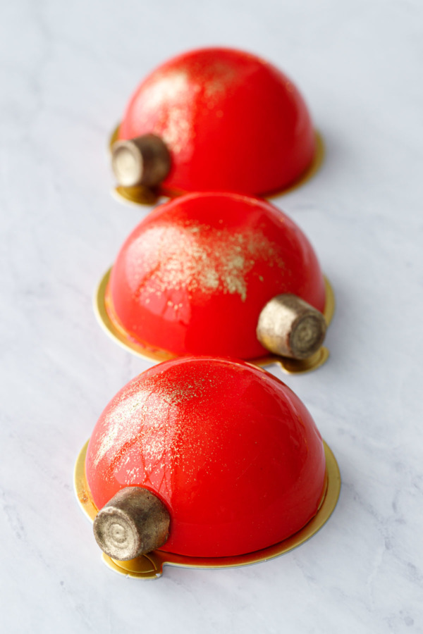 Three red mirror-glazed entremet cakes decorated with gold-dusted Rolo candies to look like Christmas ornaments