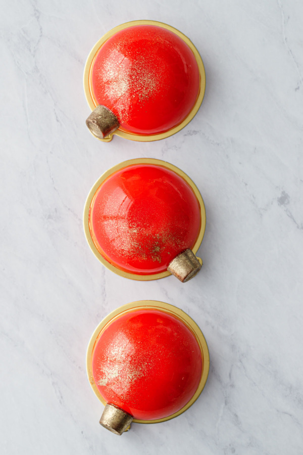 Overhead view of a row of three red mirror-glazed entremet cakes decorated with gold-dusted Rolo candies to look like Christmas ornaments