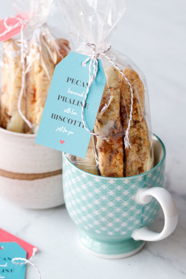 Two mugs with clear bags of biscotti cookies with red and turquoise holiday tags tied on with twine.