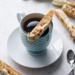 White chocolate dipped biscotti cookie sitting on the rim of a blue gingham coffee cup, with more biscotti in the background.