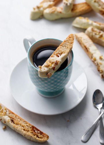 White chocolate dipped biscotti cookie sitting on the rim of a blue gingham coffee cup, with more biscotti in the background.