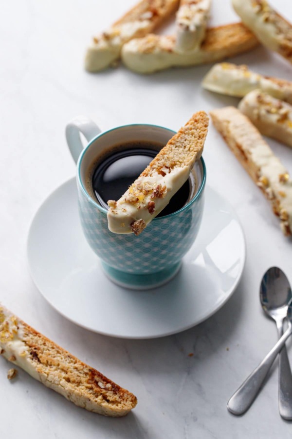 White chocolate dipped biscotti cookie sitting on the rim of a blue gingham coffee cup, with more biscotti in the background.