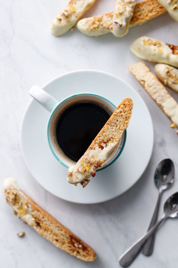 Overhead view of a biscotti cookie on the rim of a coffee cup, with two spoons and more cookies scattered about.