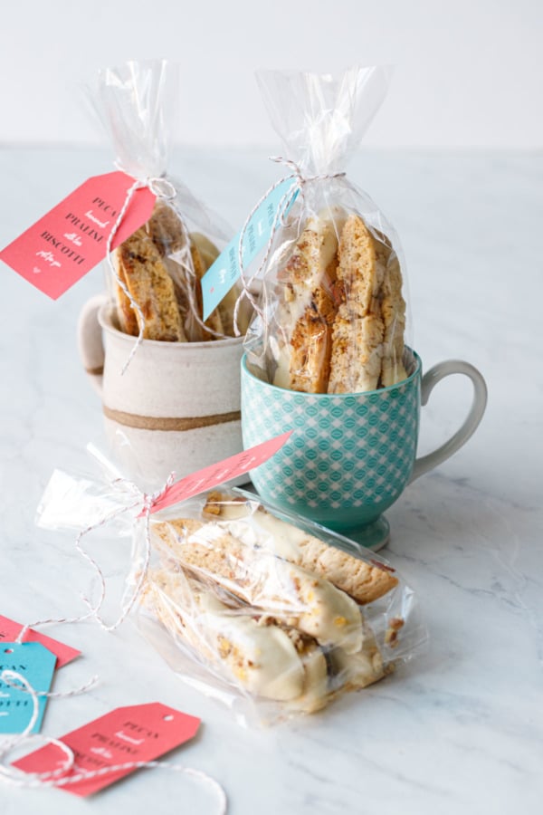 Two mugs with clear bags of biscotti cookies with red and turquoise holiday tags tied on with twine.