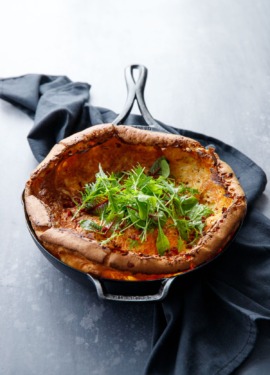Savoru Dutch baby in a Lodge cast iron skillet on a dark gray background, with a pile of baby lettuces in the middle.