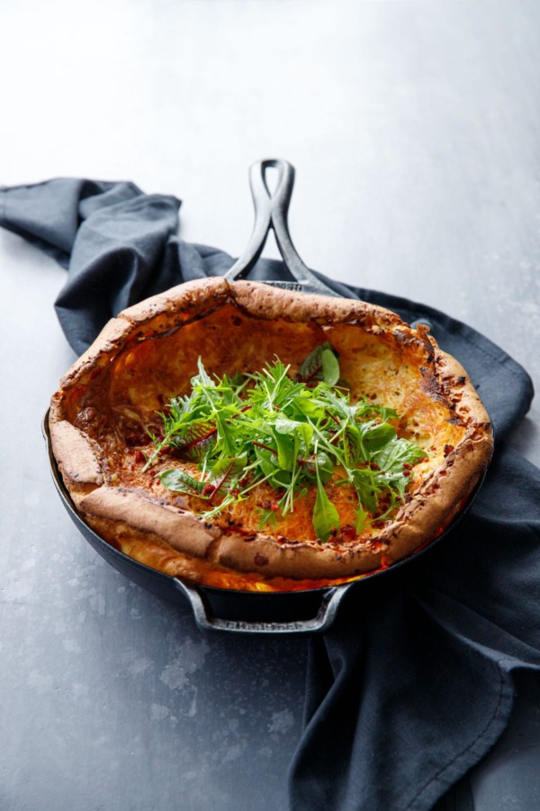 Savoru Dutch baby in a Lodge cast iron skillet on a dark gray background, with a pile of baby lettuces in the middle.