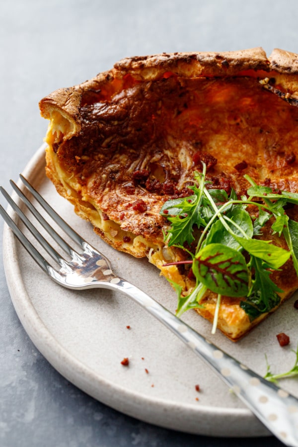 Closeup of a slice of a savory Dutch baby pancake topped with greens, on a plate with a silver fork.
