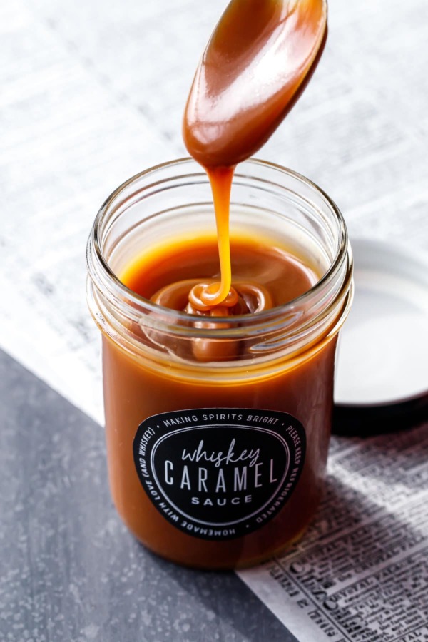 A spoon drizzling homemade whiskey caramel sauce into a jar full of caramel