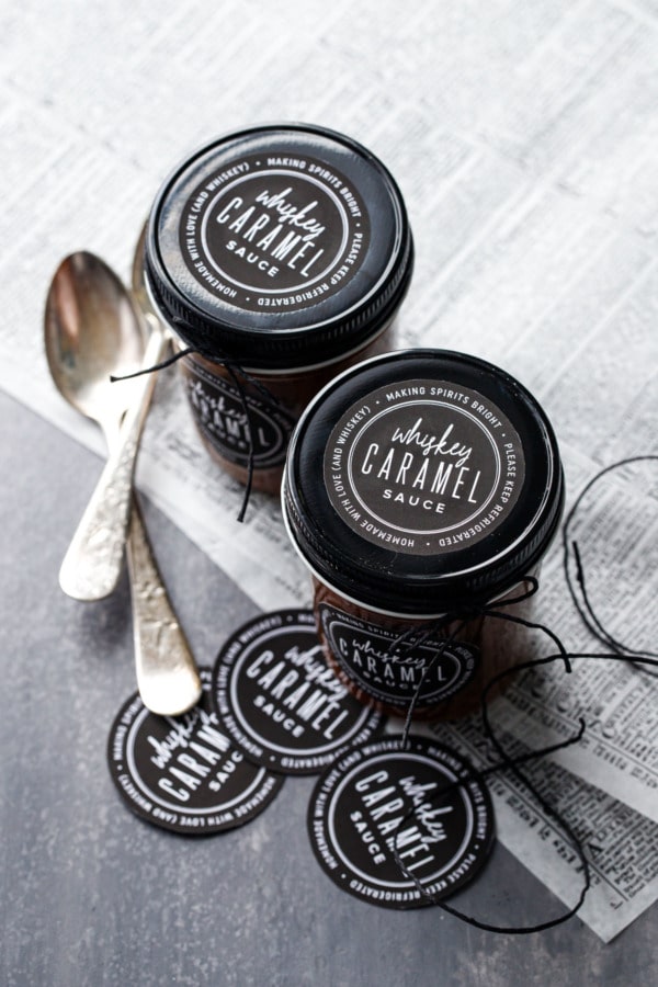 Two jars full of homemade whiskey caramel sauce, with black lids and printable black and white labels, tied with black twine.