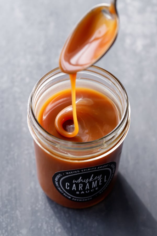 A spoon drizzling homemade whiskey caramel sauce into a jar full of caramel