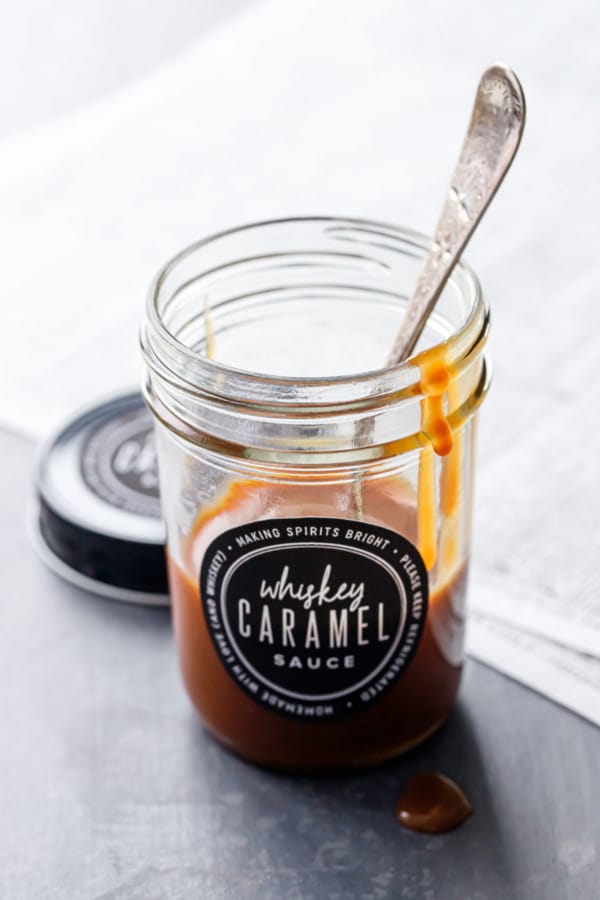 A glass mason jar partially full with homemade whiskey caramel sauce, with a few drips on the rim and a spoon in the jar.