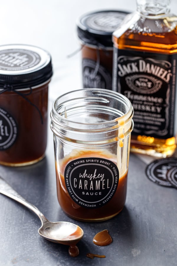 A partially full glass jar of whiskey caramel sauce, with a few more jars and a bottle of Whiskey in the background.