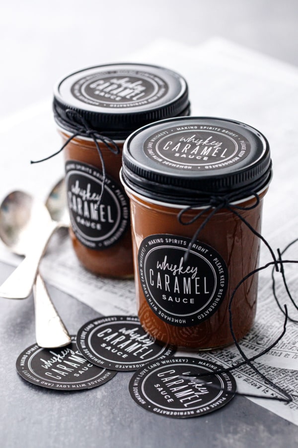 Two jars full of homemade whiskey caramel sauce, with black lids and printable black and white labels, tied with black twine.