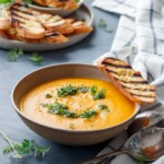 Ceramic bowl with carrot soup, topped with carrot pesto, carrot greens and a slice of grilled baguette bread, on a blue background with a plate of more bread in the background.