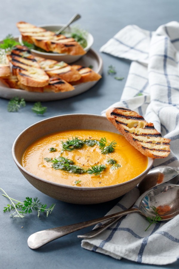 Ceramic bowl with carrot soup, topped with carrot pesto, carrot greens and a slice of grilled baguette bread, on a blue background with a plate of more bread in the background.