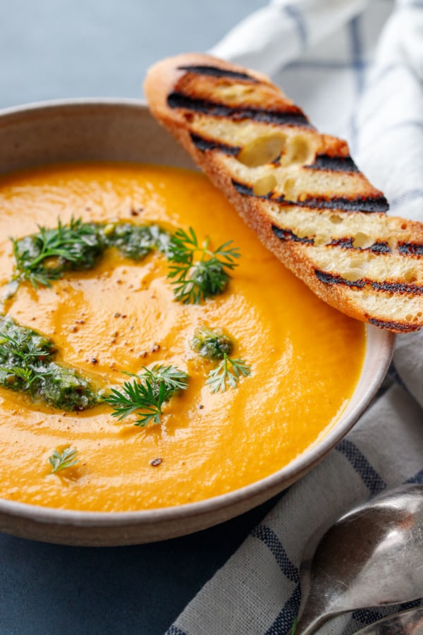 Close up shot of bowl of creamy carrot soup with dollops of carrot top pesto and a piece of grilled baguette bread resting on the rim.