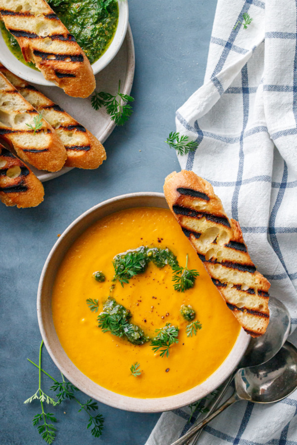 Overhead shot of bowl of creamy carrot soup with carrot top pesto and a plate of grilled baguette on the side.