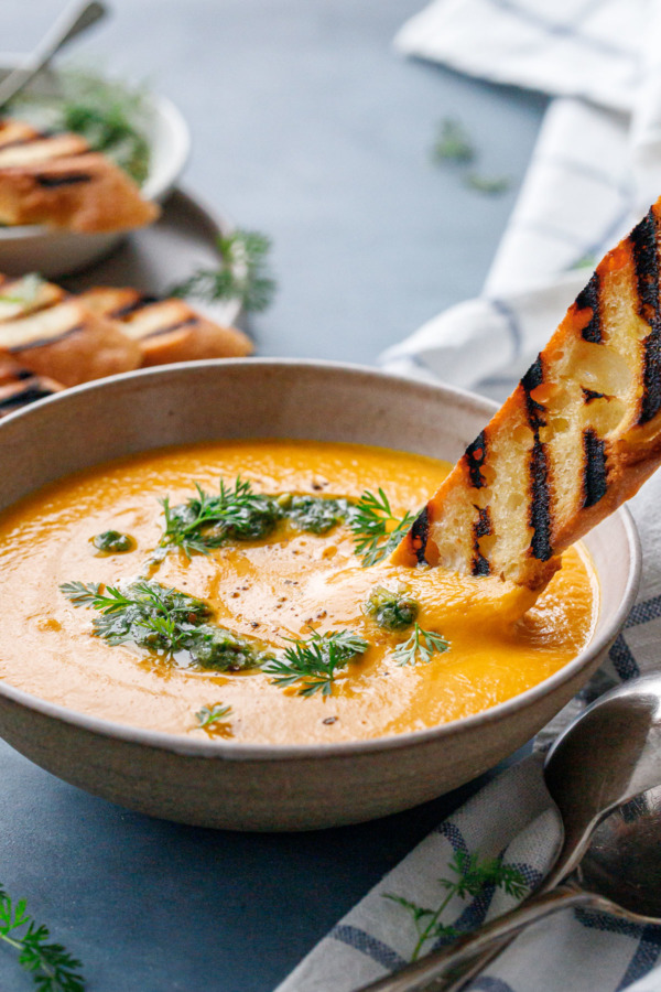 Dipping a piece of grilled baguette into a bowl of velvety carrot soup topped with carrot pesto