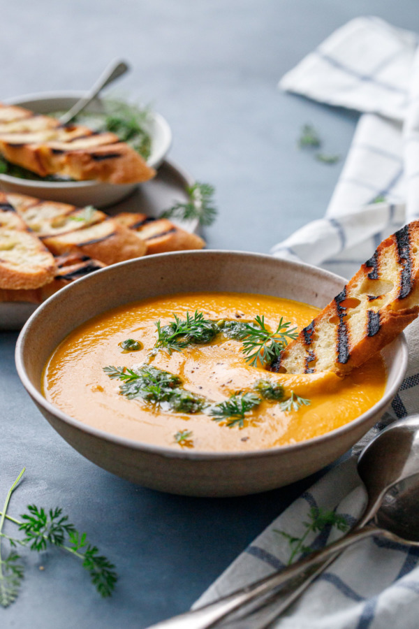 Dipping a piece of grilled baguette into a bowl of velvety carrot soup topped with carrot pesto