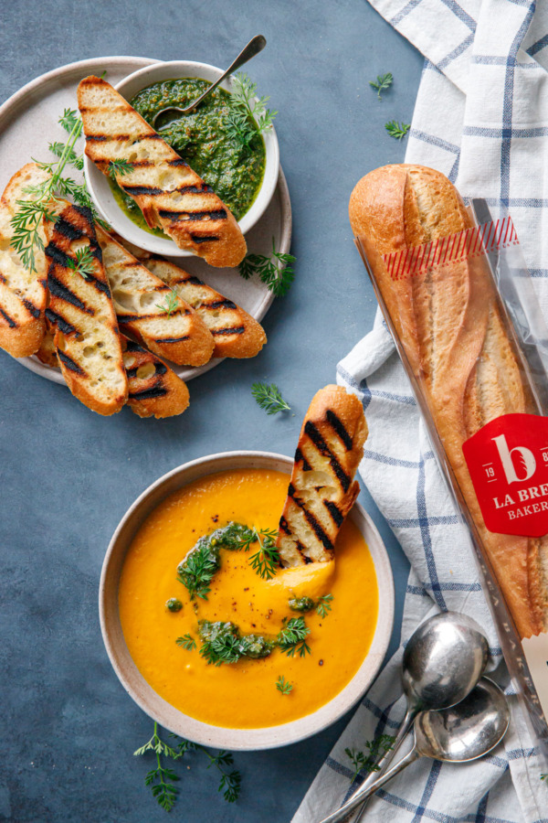 Overhead of bowl of carrot soup, plate of grilled baguette, and a full La Brea Bakery baguette in packaging.