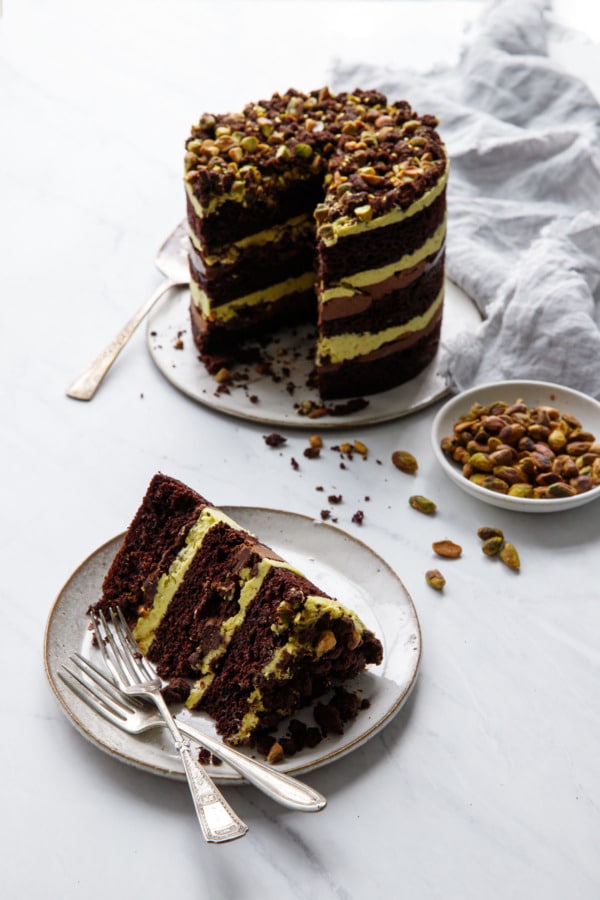 Slice of Dark Chocolate Pistachio Naked Layer Cake with the full cake in the background and a bowl of pistachios.