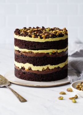 Dark Chocolate Pistachio Naked Layer Cake on a white background with a bowl of pistachios and cake server.