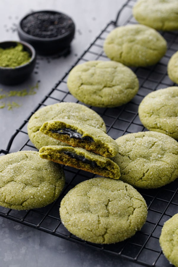 Cooling rack with rows of matcha sugar cookies, one broken in half to show black sesame filling, bowls of matcha powder and black sesame seeds.