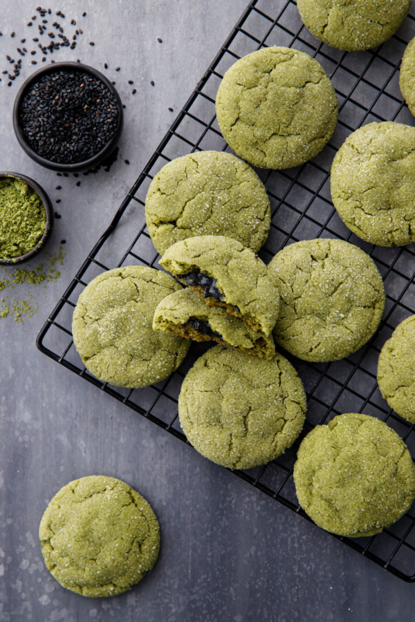 Cooling rack with rows of matcha sugar cookies, one broken in half to show black sesame filling, bowls of matcha powder and black sesame seeds.