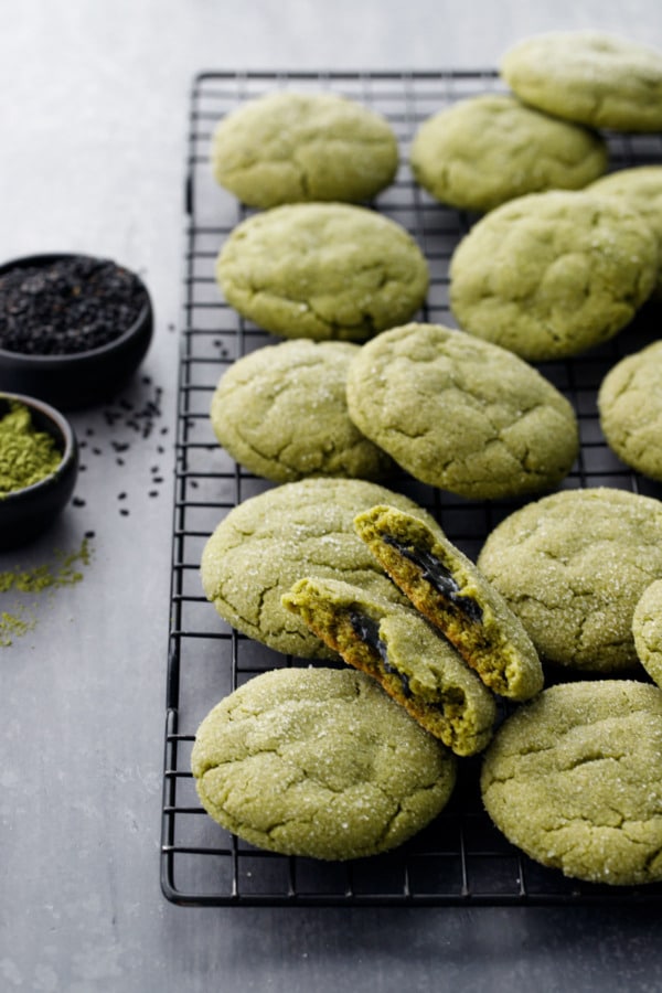Cooling rack with rows of matcha sugar cookies, one broken in half to show black sesame filling, bowls of matcha powder and black sesame seeds.