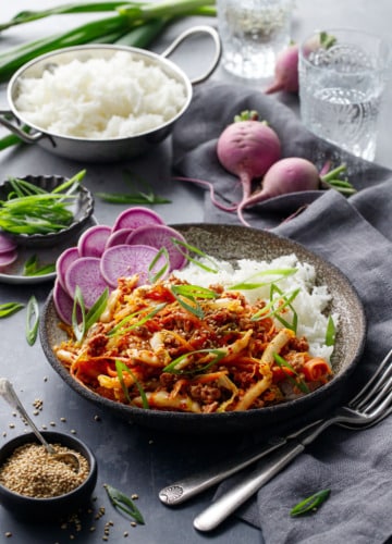 Spicy Korean Egg Roll Bowls with a bowl of rice, pinch bowl of sesame seeds, and whole purple radishes on a dark background.