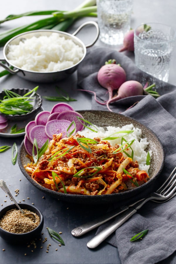 Spicy Korean Egg Roll Bowls with a bowl of rice, pinch bowl of sesame seeds, and whole purple radishes on a dark background.