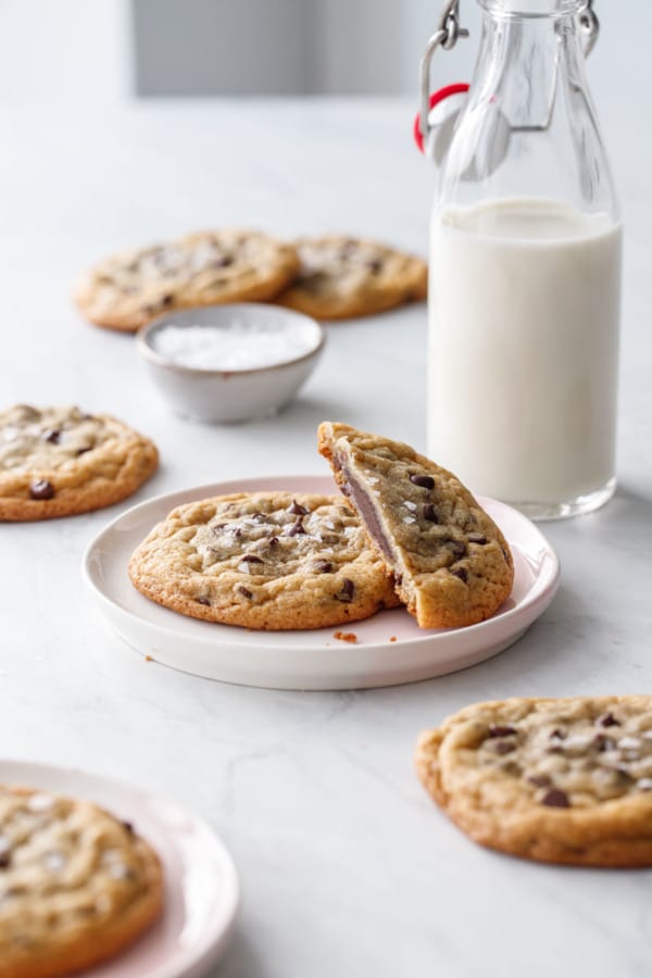 Ganache-Stuffed Chocolate Chip Cookies scattered on small plates and on marble background, with a bowl of flake salt and a milk bottle.