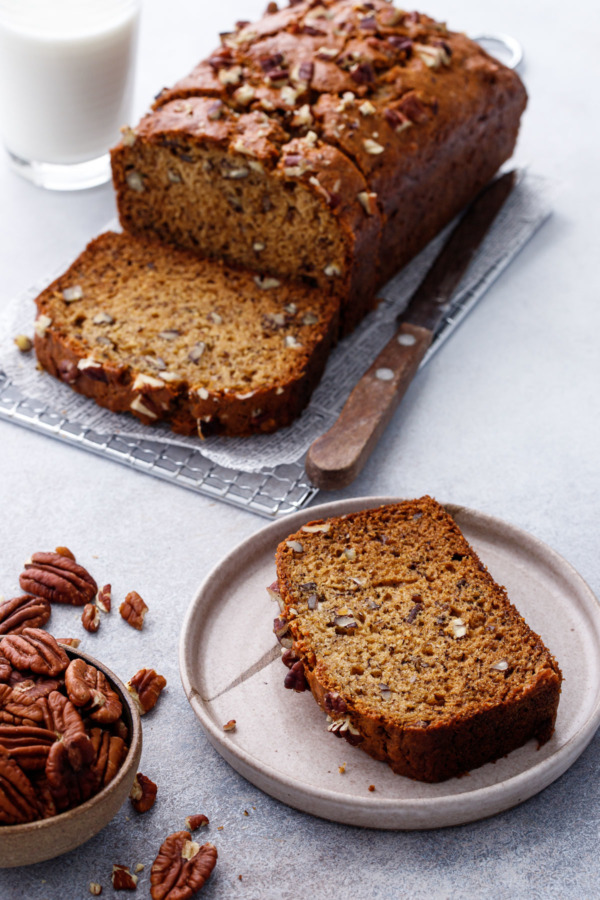 Slice of Bananas Foster Banana Bread on a plate with a bowl of chopped pecans and a glass of milk.