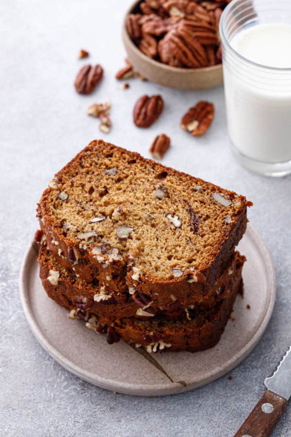 Stack of Bananas Foster Banana Bread slices on a ceramic plate, with glass of milk and bowl of pecans