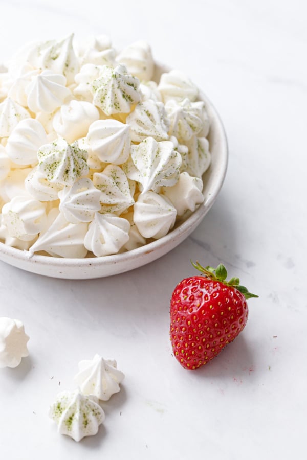 Small bowl of tiny white meringue kisses dusted with matcha, and a single strawberry