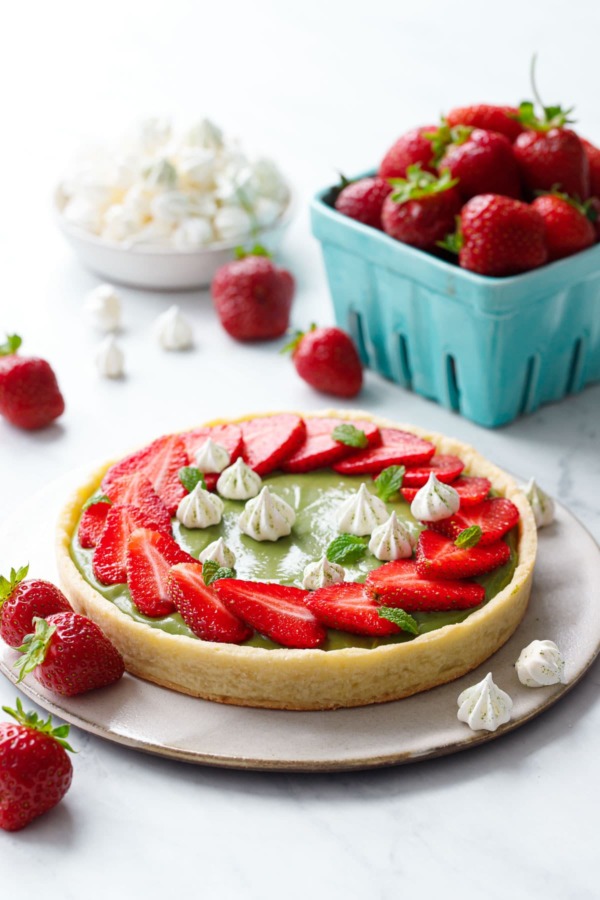 Matcha Strawberry Tart on a marble background, with turquoise basket of strawberries and a bowl of mini meringue kisses.